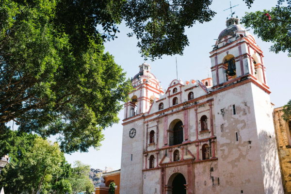 Oaxaca 2019_A historic colonial-style church with twin bell towers, faded red and white details, and surrounded by lush green trees under a clear blue sky.