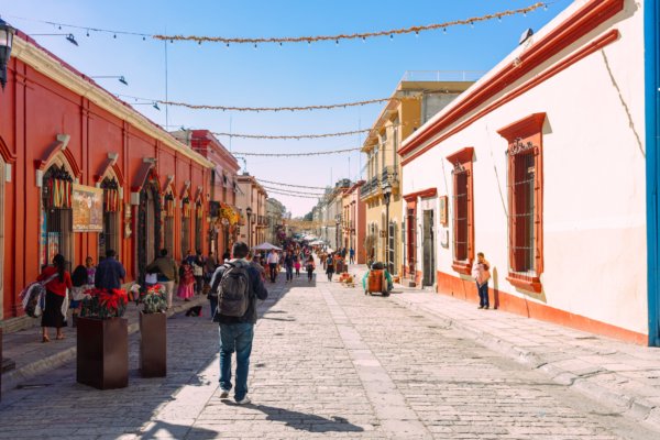 A vibrant cobblestone pedestrian street in the historic center of Oaxaca City, Mexico, lined with colorful colonial buildings in red and yellow under a clear blue sky.
