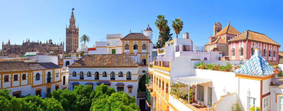 Spain 2020_Panoramic view of Seville’s historic city center with colorful Andalusian rooftops, palm trees, and the iconic La Giralda bell tower rising above the skyline under a bright blue sky.