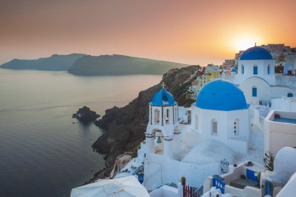 A golden hour view of the iconic blue-domed churches and whitewashed buildings of Oia, Santorini. The sun sets over the Santorini caldera and the Aegean Sea, illuminating the rugged volcanic cliffs and traditional Cycladic architecture in a warm, orange glow.