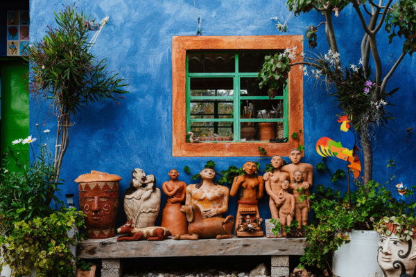 A bright blue exterior wall of an artisan studio in Oaxaca, Mexico, featuring a green-framed window and a collection of handmade terracotta sculptures, pottery, and a colorful wooden rooster outside.
