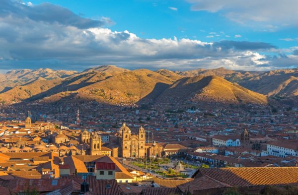 An elevated cityscape view of Cusco, Peru, showing the red-tiled roofs and historic Spanish colonial architecture of the city center, centered on the Plaza de Armas and the Cusco Cathedral, with the rolling brown peaks of the Andes Mountains under a blue sky with white clouds.