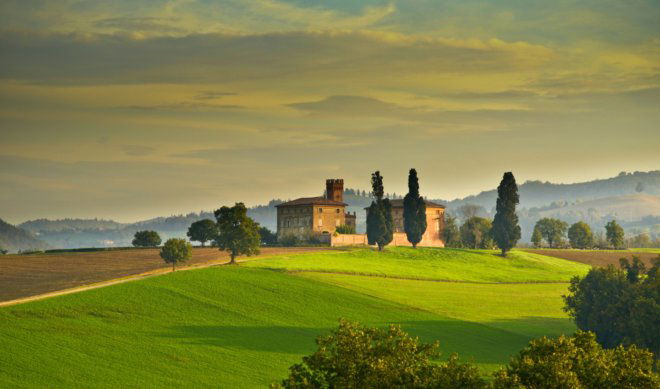 A scenic landscape of a traditional stone villa and tower nestled in the rolling green hills of Tuscany, near Florence and Siena. The image features iconic cypress trees, golden sunset light over cultivated fields, and misty Apennine mountains in the distance.