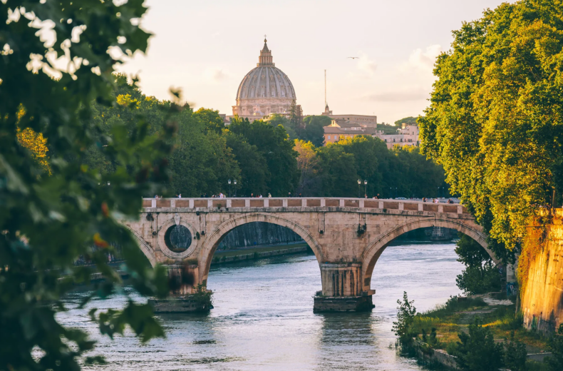 A golden hour view of the historic Ponte Sisto bridge spanning the Tiber River in Rome, with the iconic dome of St. Peter's Basilica in the Vatican City skyline. Lush green trees frame the riverbank under a soft sunset sky, capturing the timeless beauty of Italy.