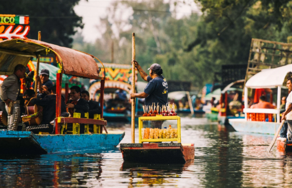 Floating snack vendor poling a small boat past colorful trajineras and mariachi musicians on the Xochimilco canals in Mexico City