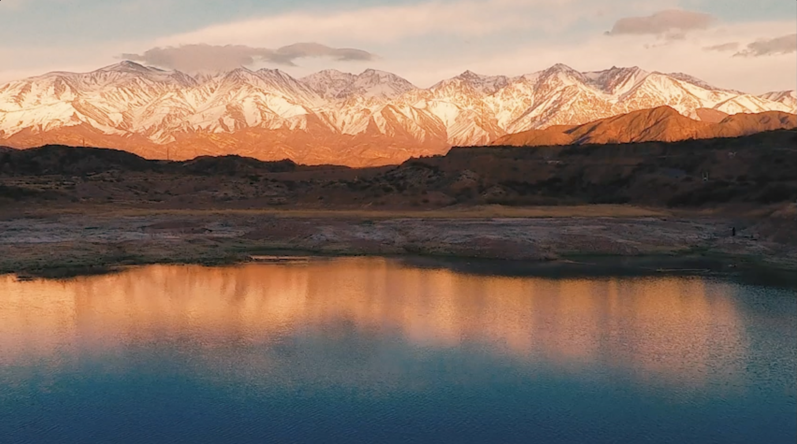A panoramic view of snow-capped mountain peaks, such as the Andes in Argentina or Chile, glowing orange and gold during sunset. The rugged mountain range is reflected perfectly in the still, blue waters of a high-altitude lake, capturing the vast, untouched wilderness of Patagonia at dusk.