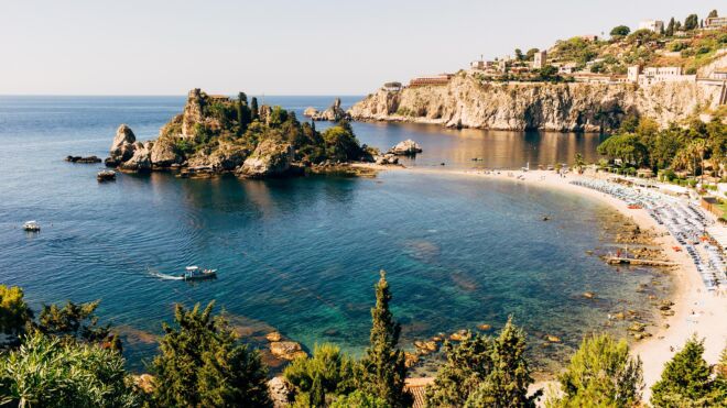 An elevated panoramic view of Isola Bella in Taormina, Sicily, featuring the small rocky island connected to the mainland by a sandy beach, surrounded by the turquoise waters of the Ionian Sea.