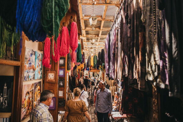 Colorful bundles of dyed wool hanging above a narrow, sunlit alleyway in the Marrakech souks, with travelers exploring local artisan shops and galleries in the heart of Morocco.