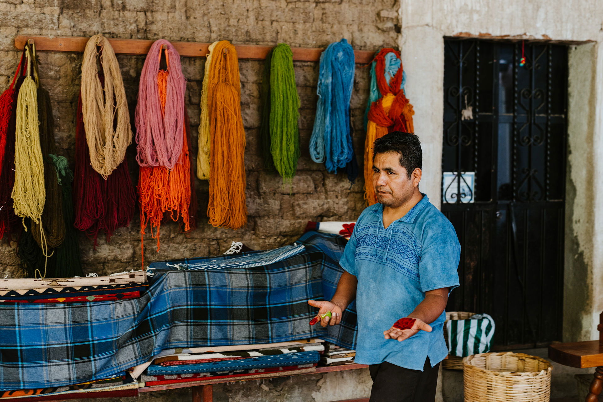 An indigenous artisan in Teotitlán del Valle, near Oaxaca City, Mexico, displays natural pigments in his hands. Behind him, vibrant skeins of hand-dyed wool in blue, gold, and green hang against an adobe wall, showcasing the traditional textile craftsmanship of the region.