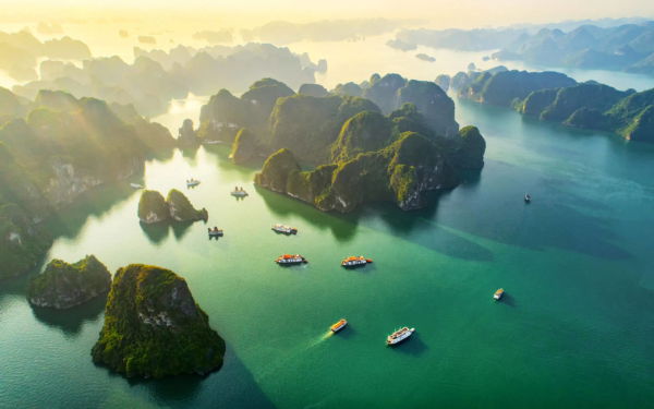 Boats scattered across the turquoise waters of Hạ Long Bay at sunrise, surrounded by misty limestone islands.