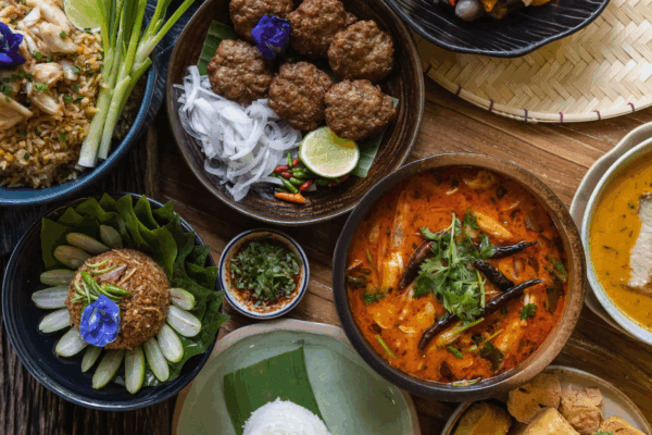 Thailand 2026_ A colorful spread of traditional Thai cuisine, including spicy red curry, fried rice, fresh vegetables, meat patties, and dipping sauces, beautifully arranged on a wooden table with banana leaves and edible flowers.