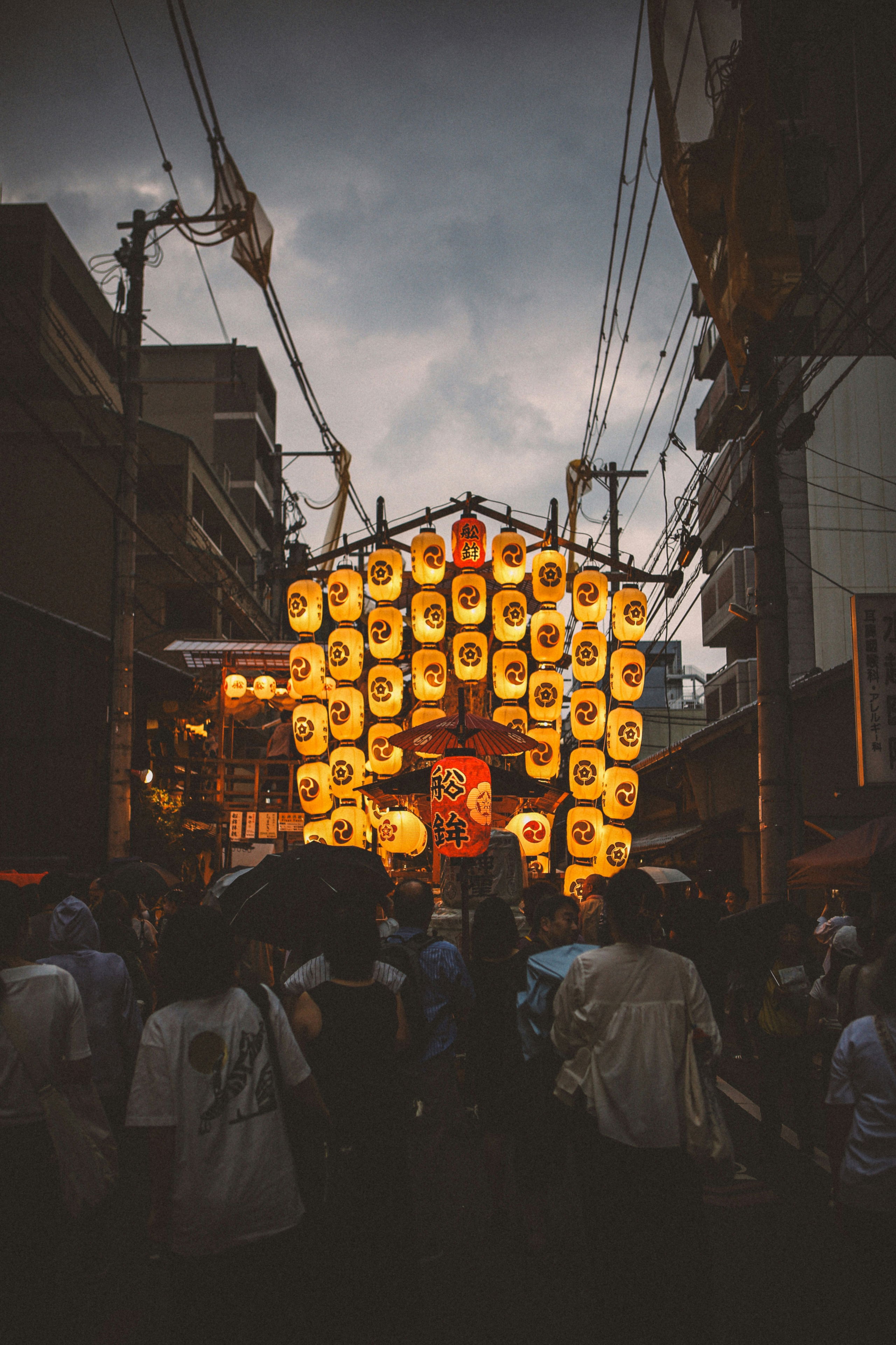 A crowd gathers beneath glowing lanterns and wooden floats in downtown Kyoto as dusk falls during Gion Matsuri’s vibrant Yoiyama evening celebrations.