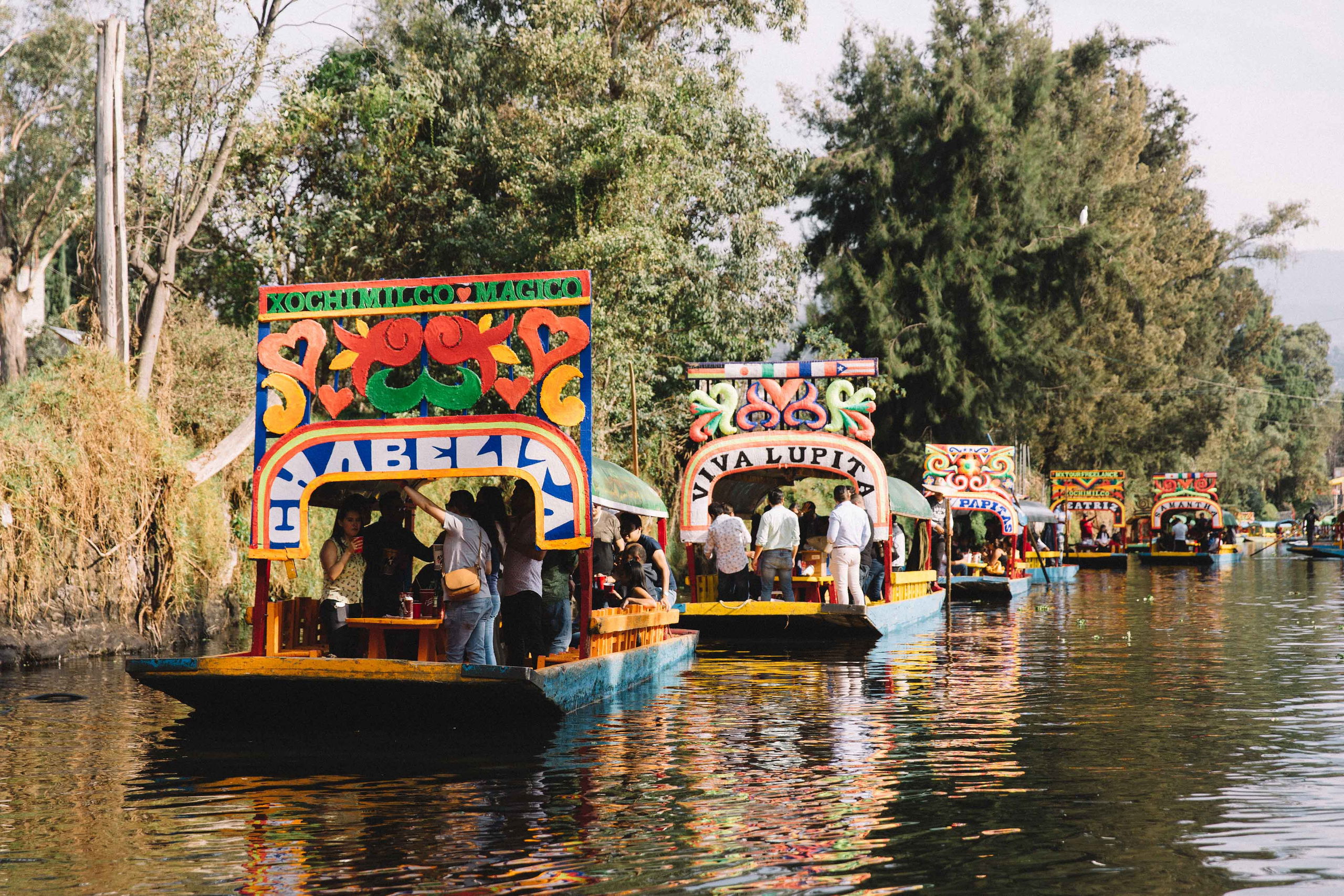 Brightly painted trajineras float along the canals of Xochimilco, Mexico City, as locals and visitors enjoy a festive day surrounded by music, food, and vibrant scenery.