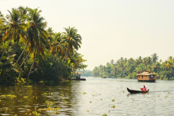 India 2025_Serenely gliding through the Kerala backwaters, a small canoe passes lush palm-lined shores and traditional houseboats, capturing the tranquil rhythm of life in southern India.